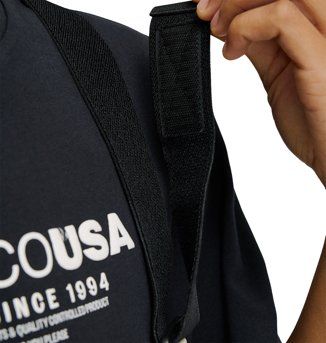 Wearing a dark shirt with white text partially reading “COUSA SINCE 1994,” a person adjusts the black strap of the Mountain Lab Bib 20K from DC Shoes, ready for any adventure with breathable protection.