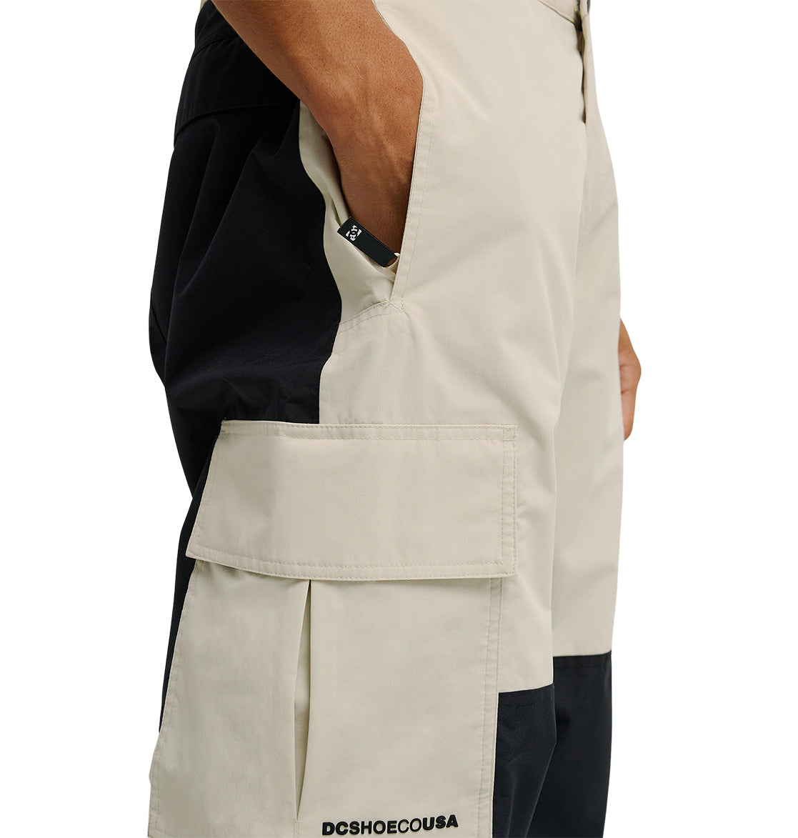 A person wearing DC Shoes Mountain Lab Pants 10K in a two-tone black and beige design, featuring a large side pocket with their hand inside. These pants are designed for breathable protection and comfort and have “DCSHOECOUSA” printed on them.