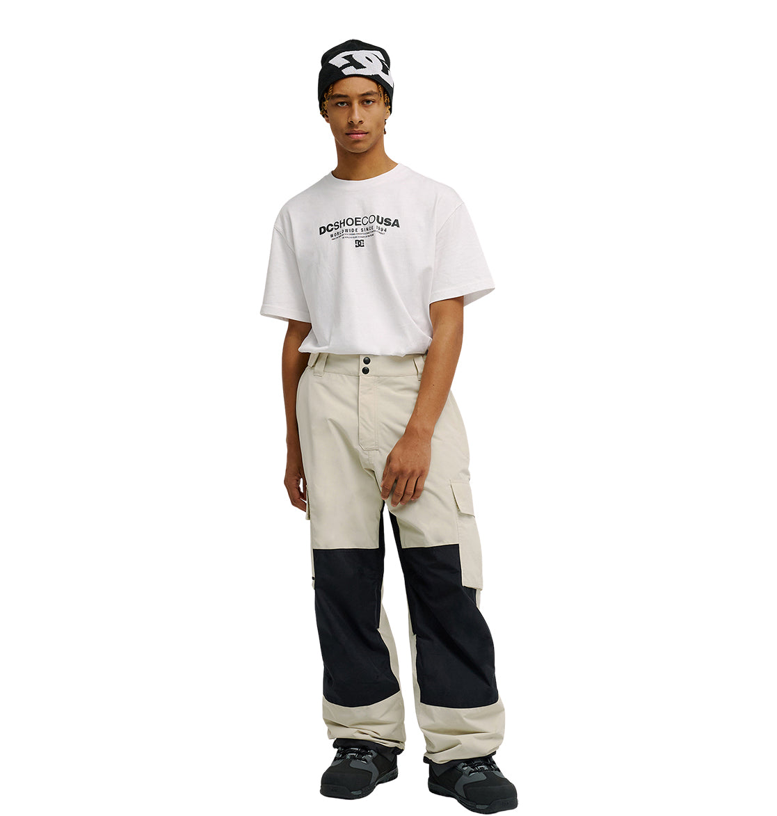 Wearing a black-and-white headband, a white DC SHOES USA t-shirt, Mountain Lab Pants 10K by DC Shoes—beige cargo pants with black knee patches featuring breathable protection—and black sneakers, the person stands against a plain white background.