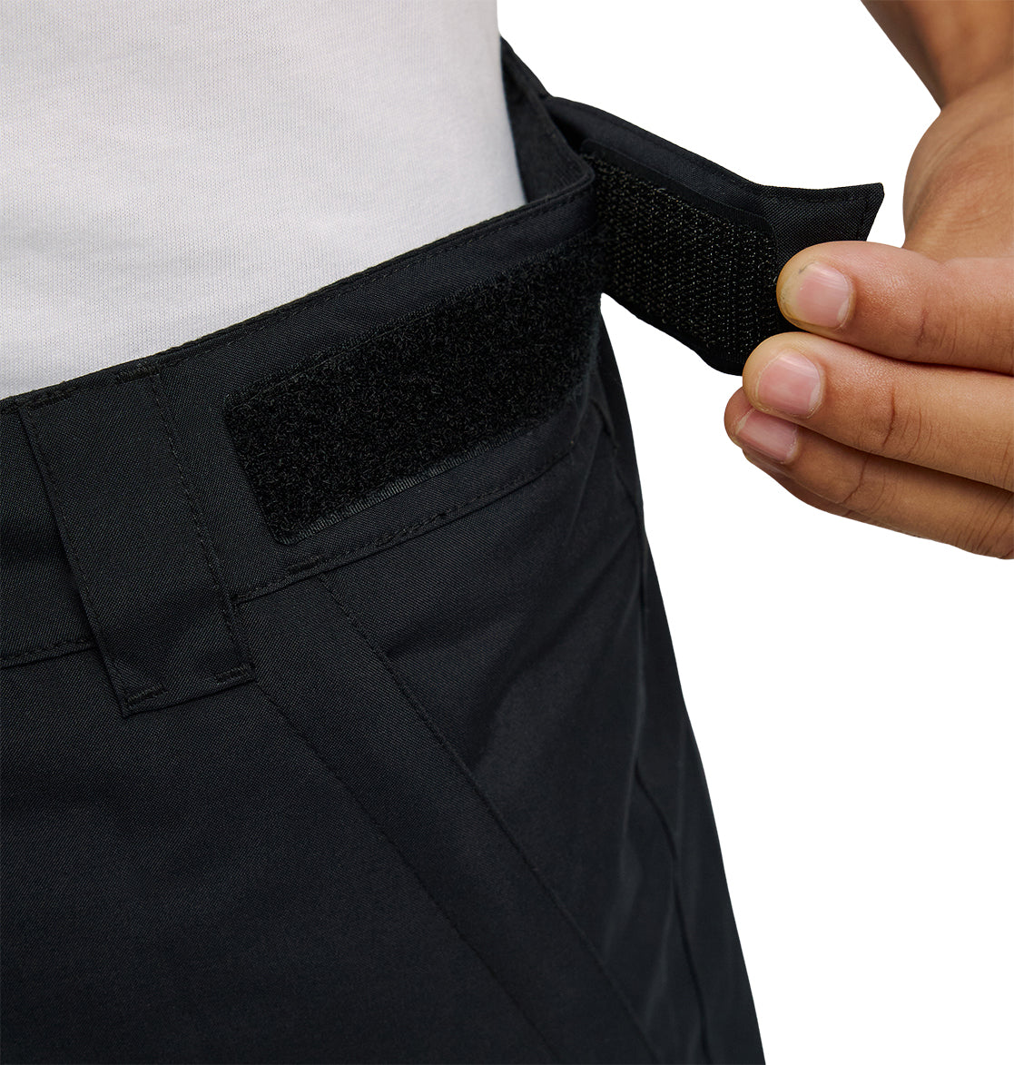 A hand adjusts the black Velcro waistband on a pair of DC Shoes Banshee Pants, which are waterproof snow pants. The person is wearing a white shirt.