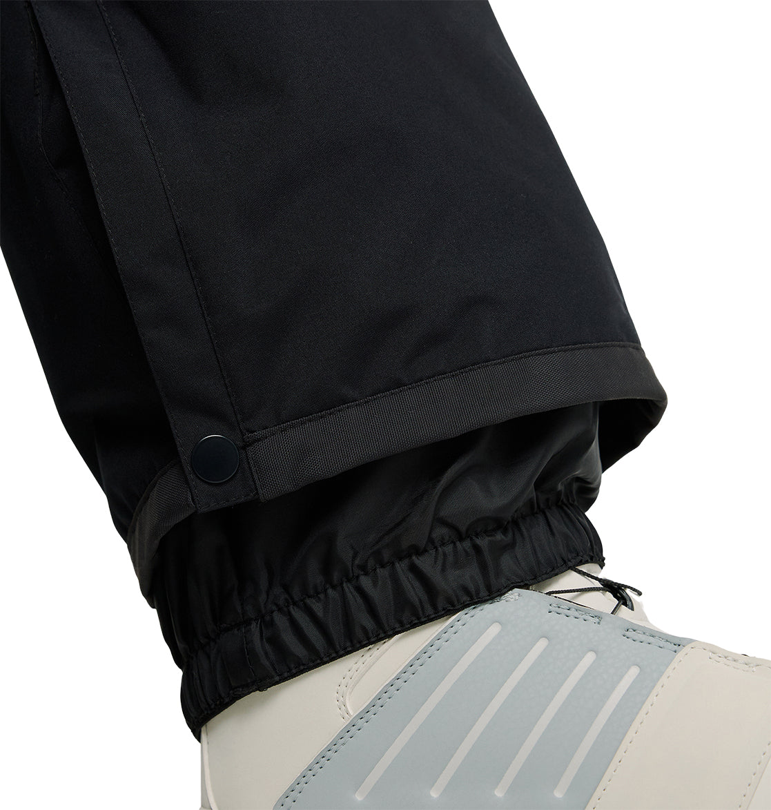 Close-up of a person wearing DC Shoes Banshee Pants, featuring black insulated snow pants with an elastic cuff and a button near the ankle for extra protection over white and gray winter boots. The Banshee Pants offer added warmth and waterproof performance in snowy conditions.