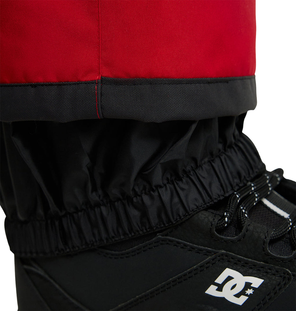 Close-up of a red and black DC Shoes Banshee Pant leg with an elastic cuff, tucked into a black winter boot featuring visible laces and part of the white DC logo.