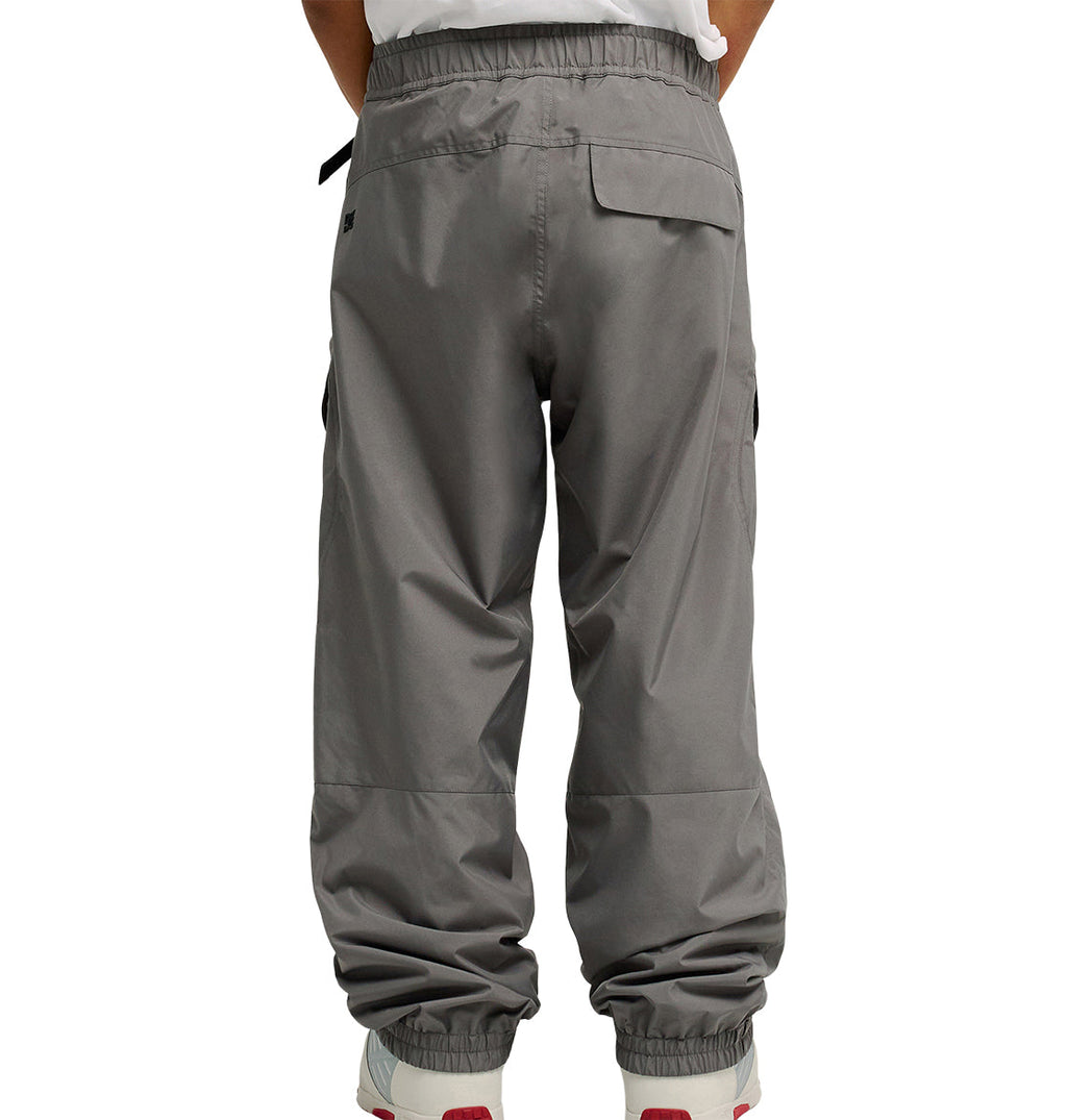 A person is shown from the back wearing DC Shoes Smoke Pants 10K in gray, along with white sneakers, standing with hands in their pockets. These pants feature an elastic waistband and elastic ankle cuffs, providing breathable protection and all-day comfort.