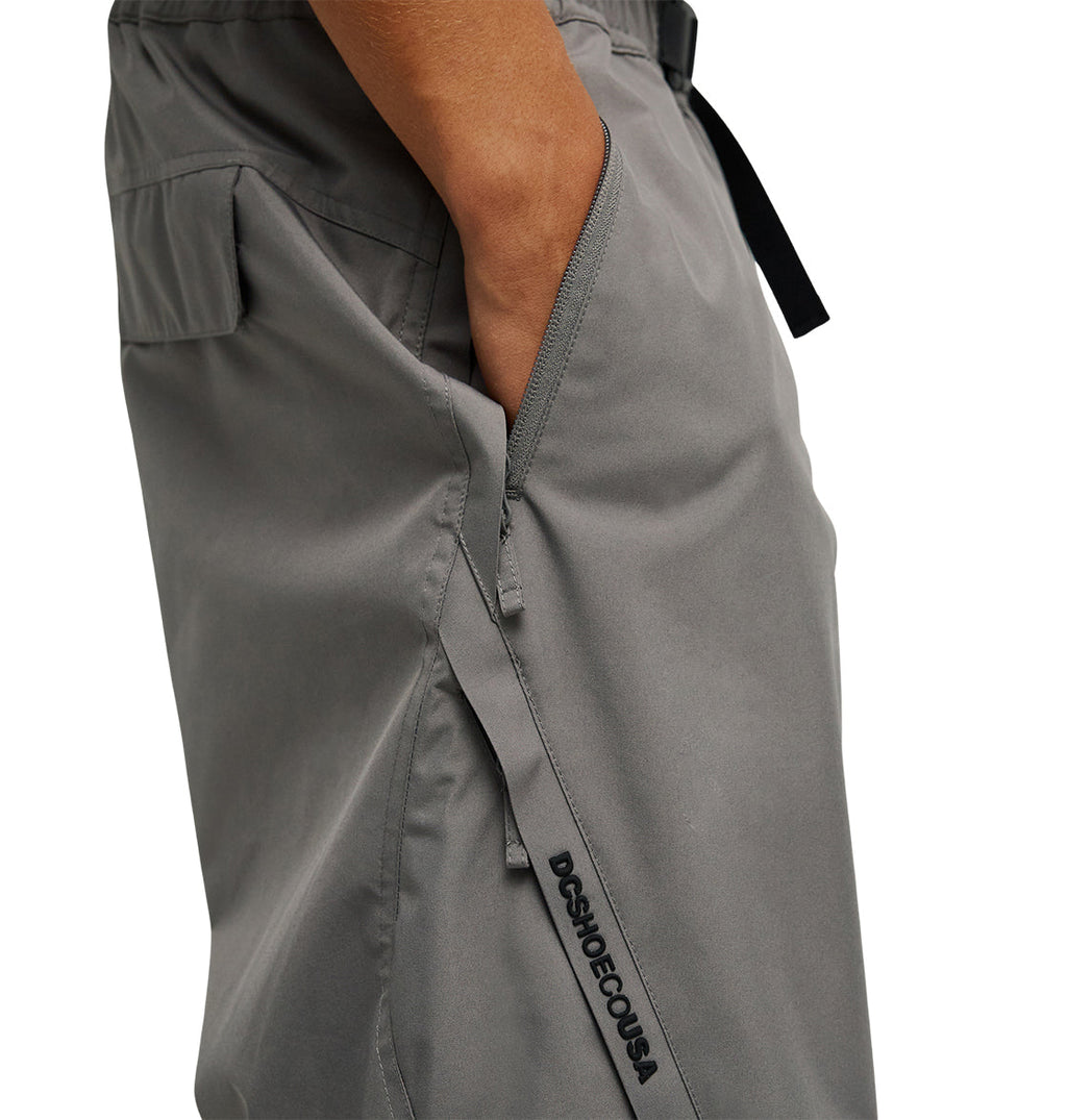 A person wearing DC Shoes Smoke Pants 10K in gray, featuring a breathable design, a black side zipper pocket, and “DSHO DECOUSA” text detailing, rests their hand in the pocket.