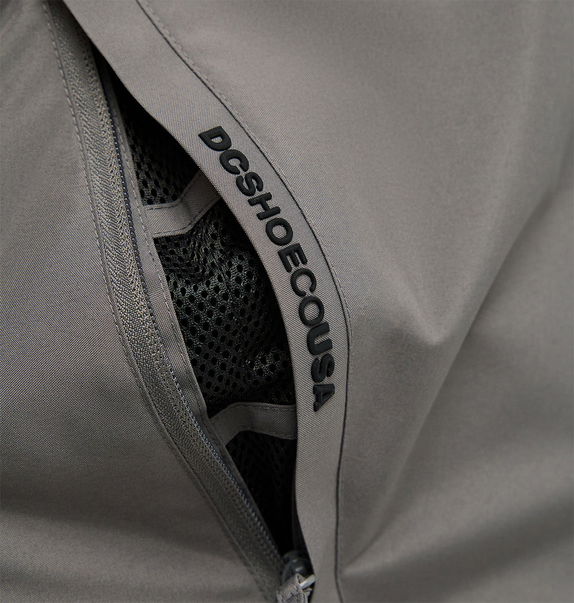 Close-up of a gray zippered pocket on the Smoke Pants 10K by DC Shoes, featuring black mesh lining for breathable comfort and “DCSHOECOUSA” printed along the zipper seam.