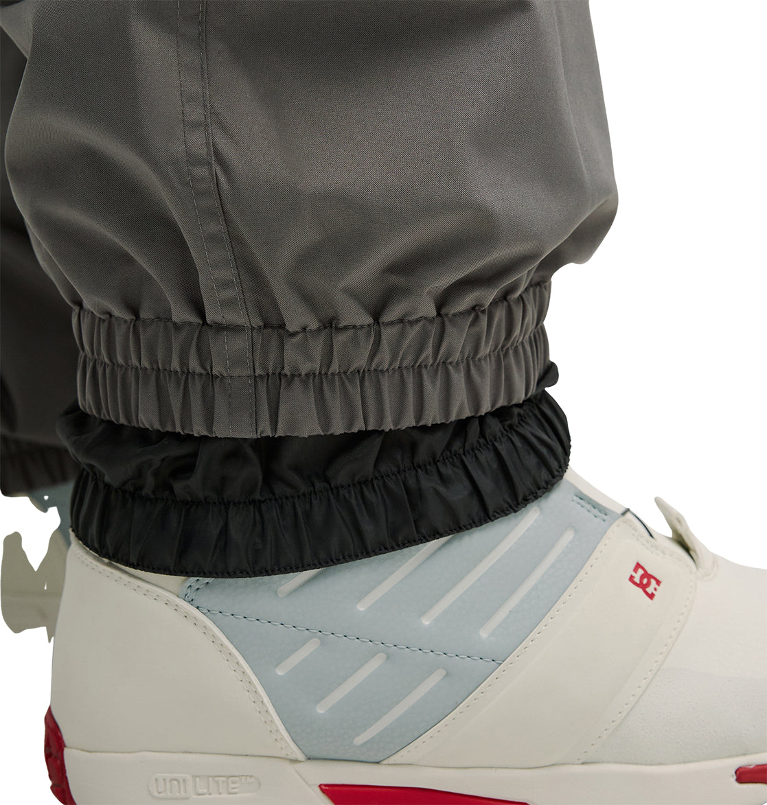 Close-up of a person wearing DC Shoes’ Smoke Pants 10K in gray, featuring elastic cuffs layered over black, paired with white sneakers with red and gray accents. These pants offer breathable protection for enhanced comfort and style.