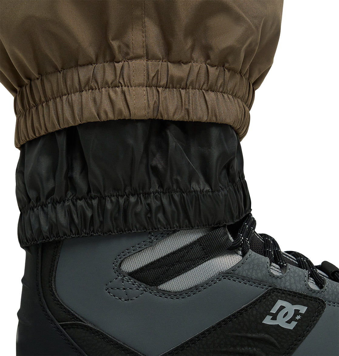 Close-up of the brown and black elastic cuffed Smoke Pants 10K by DC Shoes over a black and gray DC brand snow boot, highlighting the breathable materials and precise stitching details.