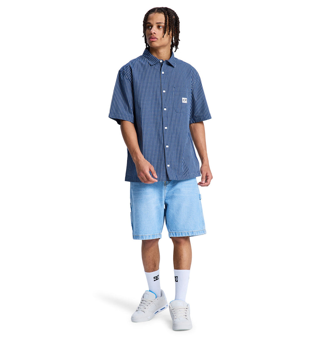 A person wearing the Reebok Game On Ss short-sleeve blue checkered button-up shirt, light blue denim shorts, white crew socks, and white sneakers stands against a plain white background.