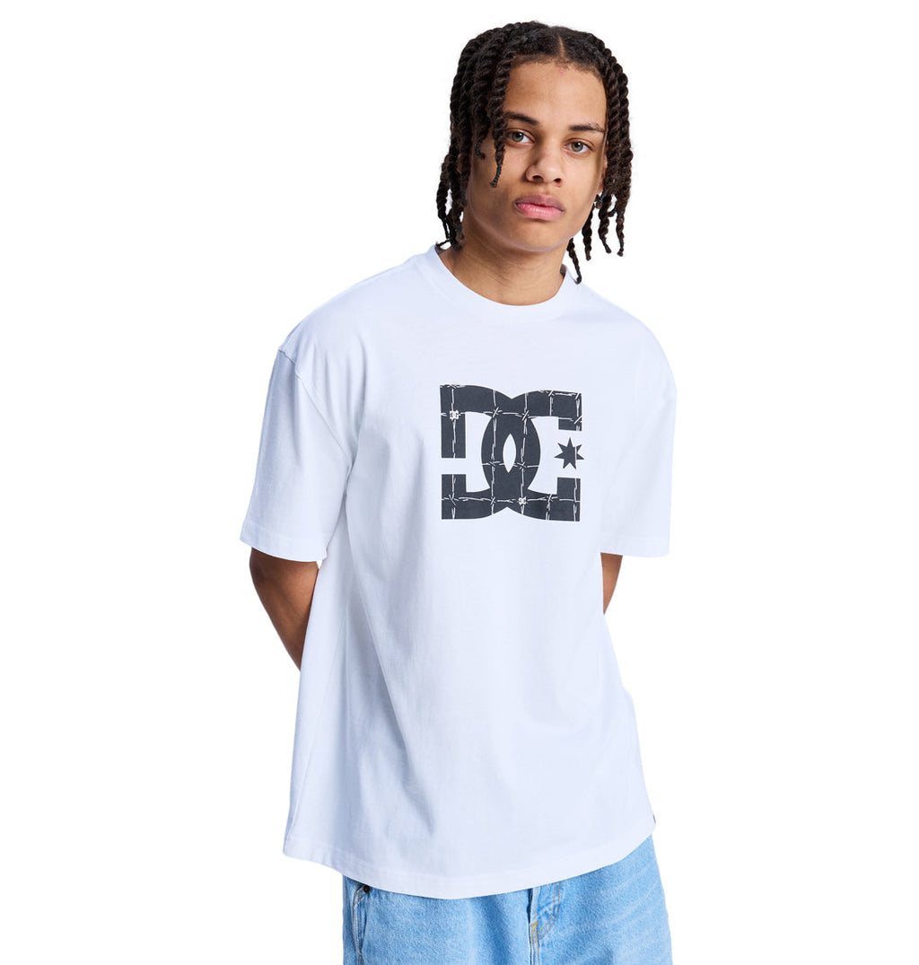 A young person with braided hair wears a white Reebok DC Star Filled Hss t-shirt featuring a large black logo on the front, paired with light blue jeans, standing against a plain white background.