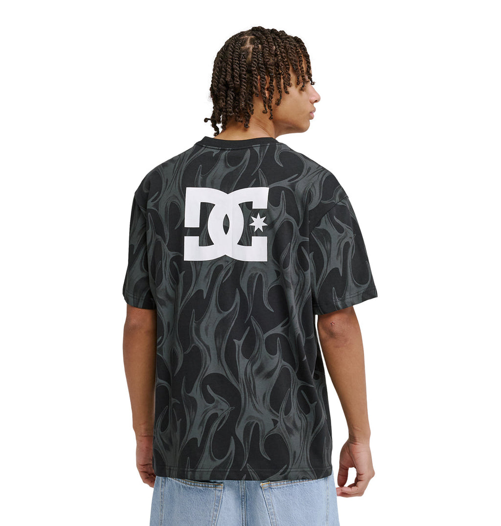 A person with braided hair wears the Reebok DC Liquid Fuego Hss T-shirt, featuring a gray flame pattern and large white logo on the back, paired with light blue jeans and facing away.
