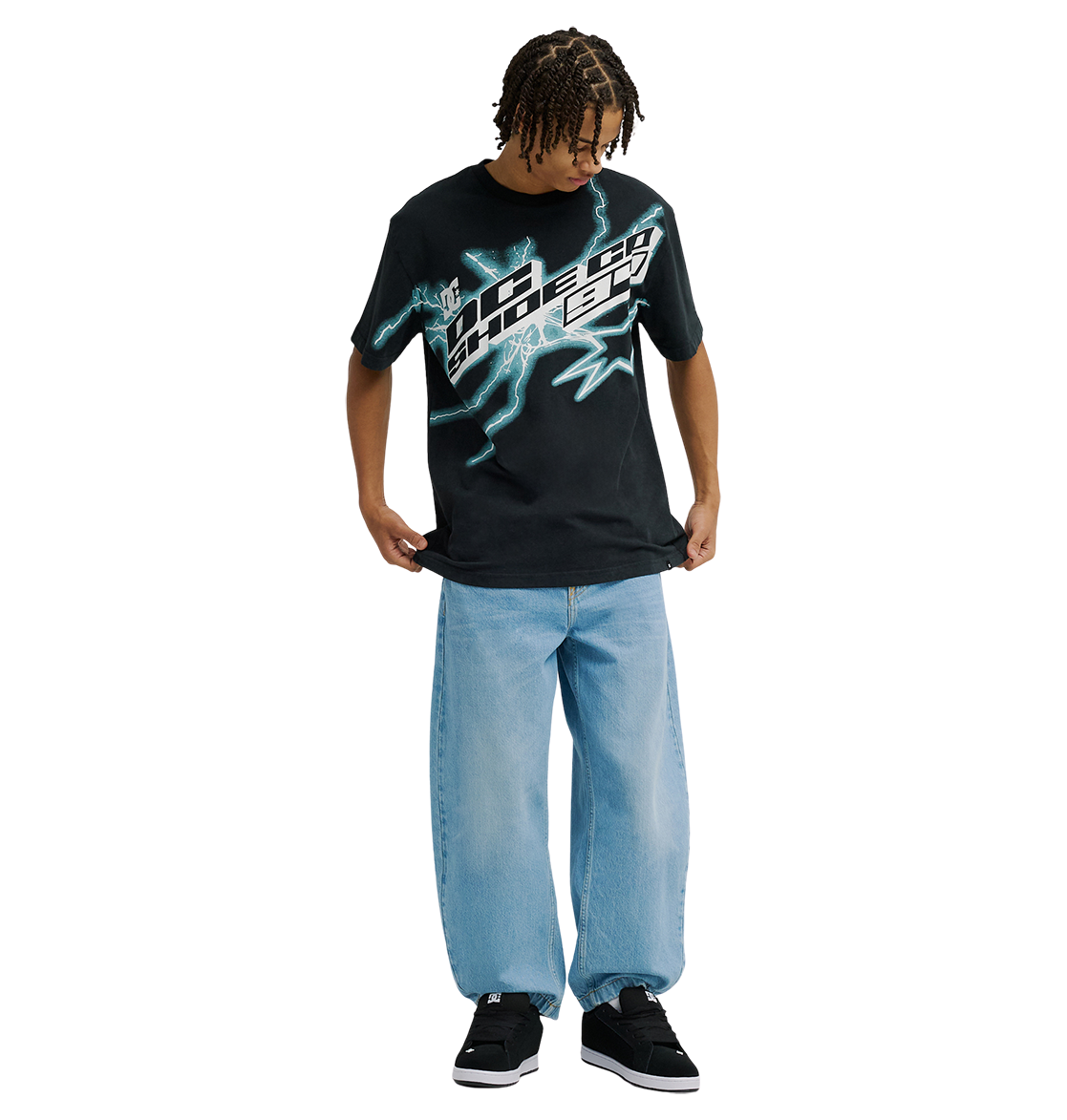 A person stands looking down, wearing the Men's DC Star Struck Tee by DC Shoes—a relaxed fit black graphic skate shirt featuring blue lightning designs—paired with light blue baggy jeans and black sneakers. The background is plain white.