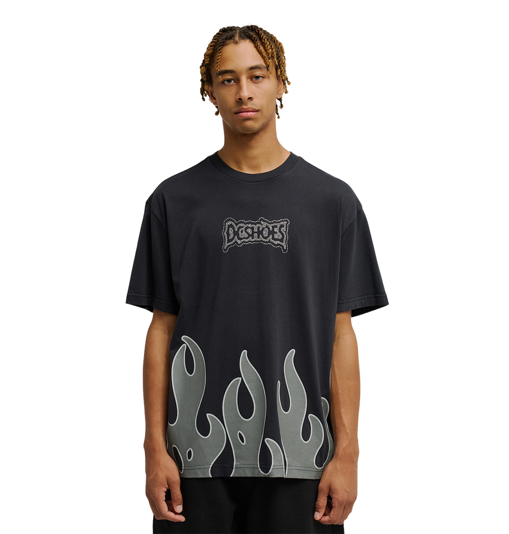 A young man with short braids wears the Men's DC Burning Diamonds Tee from DC Shoes—a black graphic skate t-shirt featuring the iconic Dogshoes logo on the chest and gray flame graphics at the hem, inspired by burning diamonds artwork. He stands out against a plain, light background, looking directly at the camera.