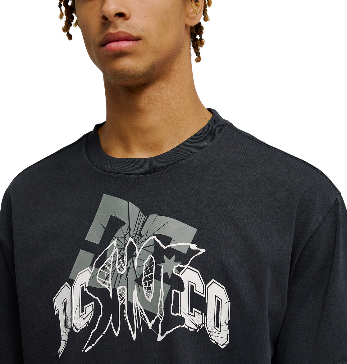 A person wearing the Men's DC Speratic Tee by DC Shoes—a black, relaxed-fit skate t-shirt featuring a bold, stylized gray and white "DC SHOCO" cracked-effect graphic. The heritage-inspired artwork stands out while only the lower half of the face is visible.
