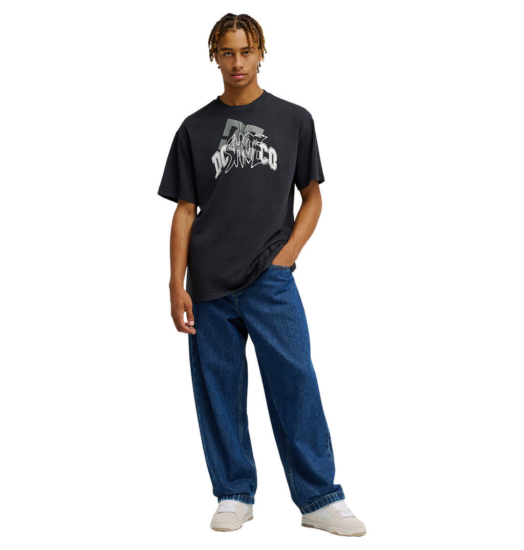 A person wearing the Men's DC Speratic Tee by DC Shoes—a black graphic skate shirt with heritage-inspired artwork—paired with loose blue jeans and white sneakers stands against a plain white background, one hand in their pocket.
